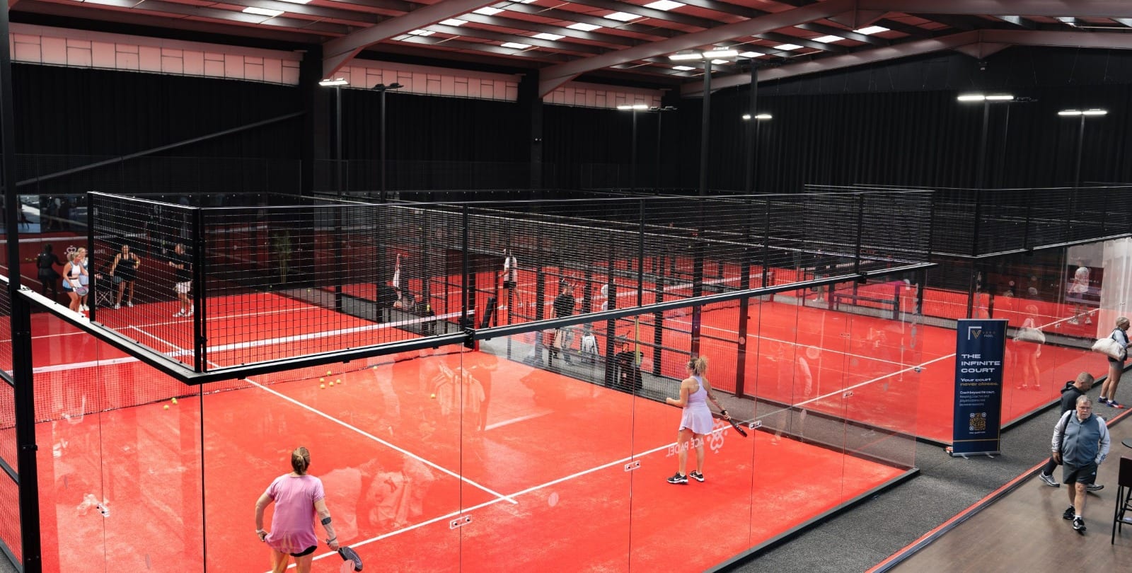Indoor padel courts at Ace Padel, Lytham, with red playing surfaces and glass-and-mesh enclosures under a steel-framed roof, players mid-game