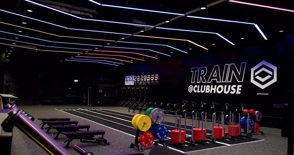 The gym at Clubhouse Blackburn: a black-walled hall under angled neon ceiling strips, with weight plates on sleds along a sprint track and Train @Clubhouse on the end wall