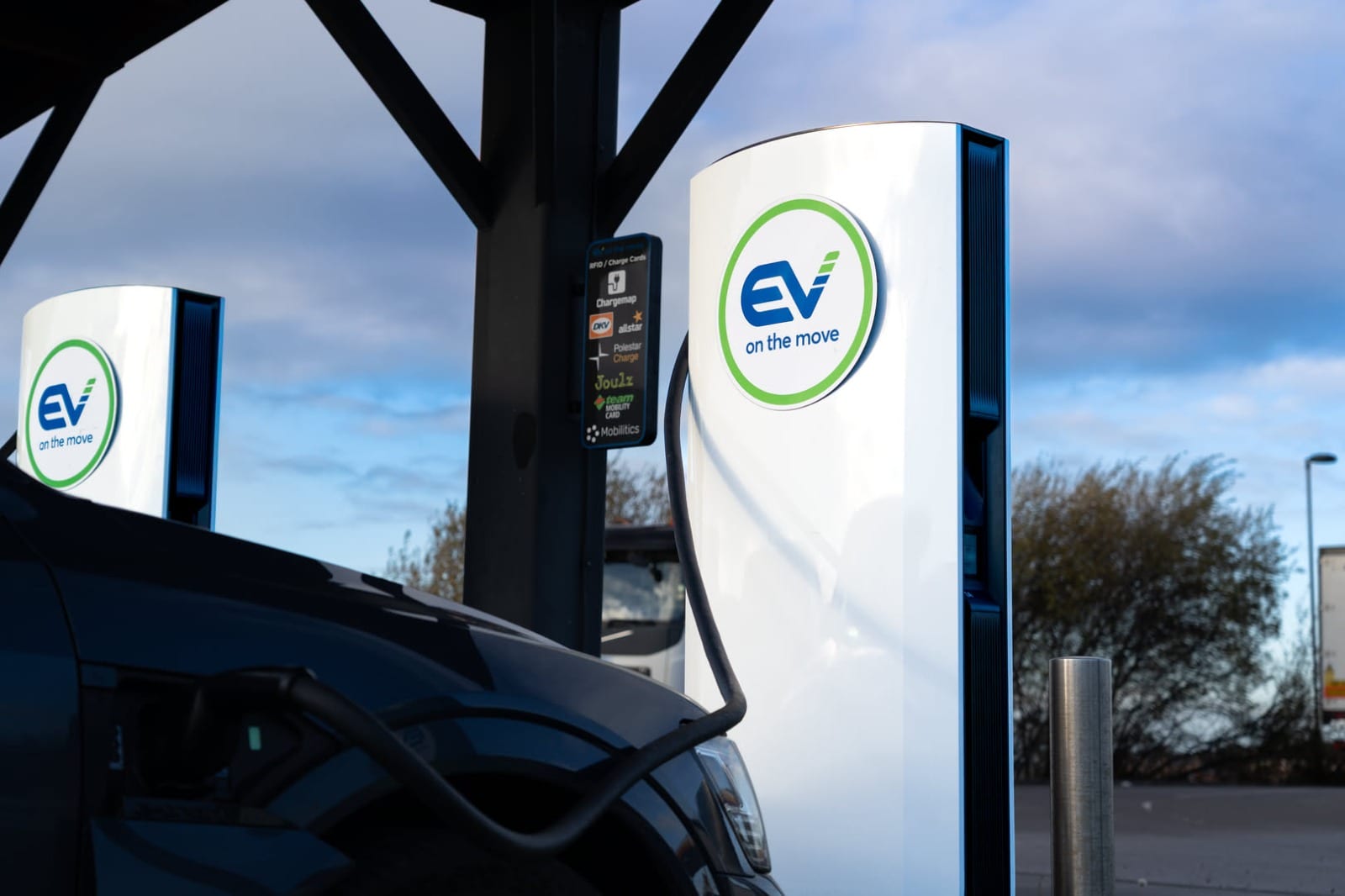 A rapid EV charger under a canopy with a car plugged in, a second charger and an HGV behind, against an evening sky